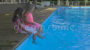 两个小女孩在游泳池里泡脚的户外镜头。 可爱的小女孩坐在游泳的边缘